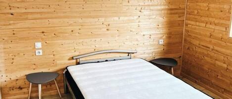 A small, single-bed room with wooden walls, a metal bed frame, a small table, and a chair.