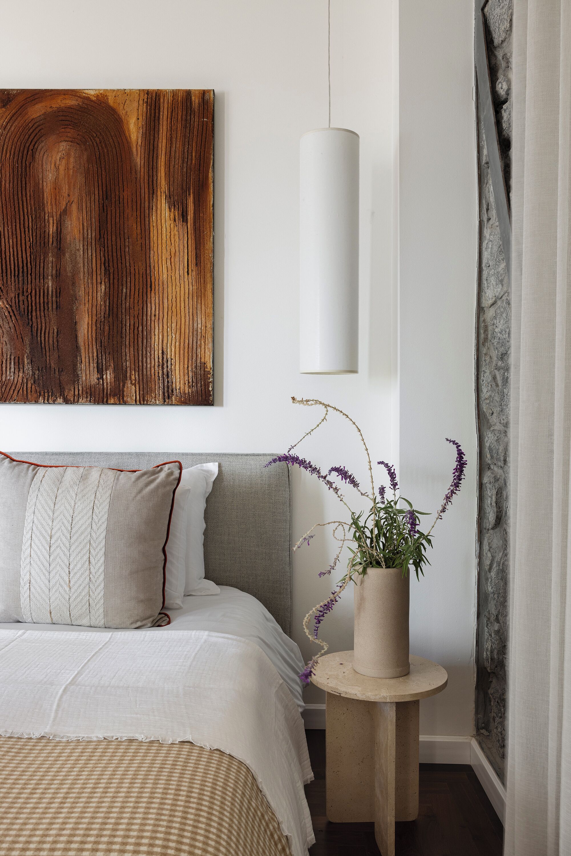 A bedroom with a bed, pillows, a bedside table with a plant, a wall-mounted light, and a large abstract painting.