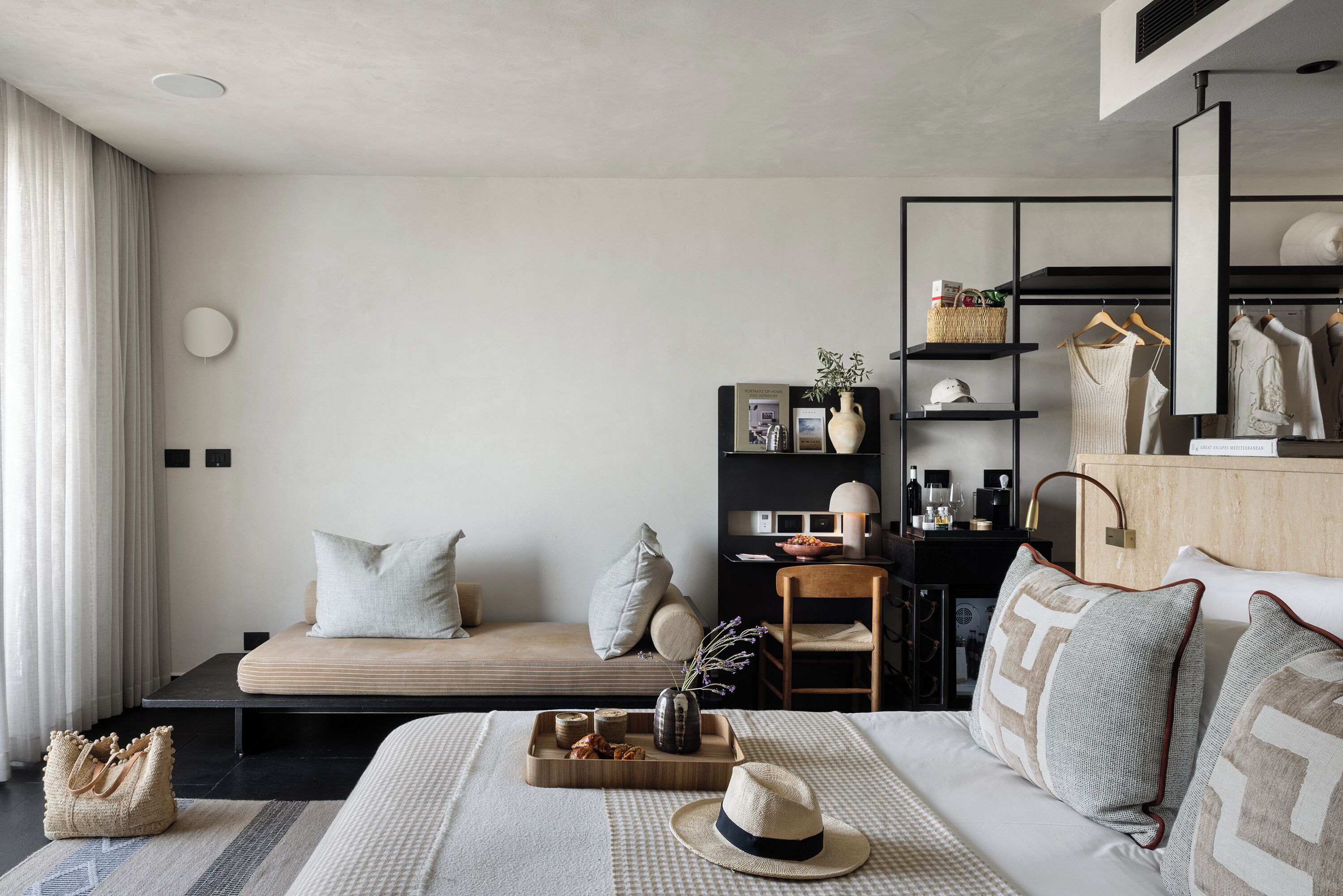 A modern hotel room with a sofa, a bed with decorative pillows, a tray with snacks, a hat, and a mirror.