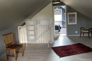 Een kamer met een schuine balkenkap, een houten stoel, een witte opbergkast, een rode vloerkleed en een deuropening naar een andere kamer met een bed en een kroonluchter.