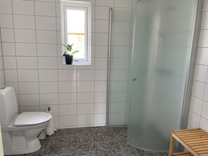 Een badkamer met witte tegels, een toilet, een douche met een matglazen deur en een klein houten krukje.
