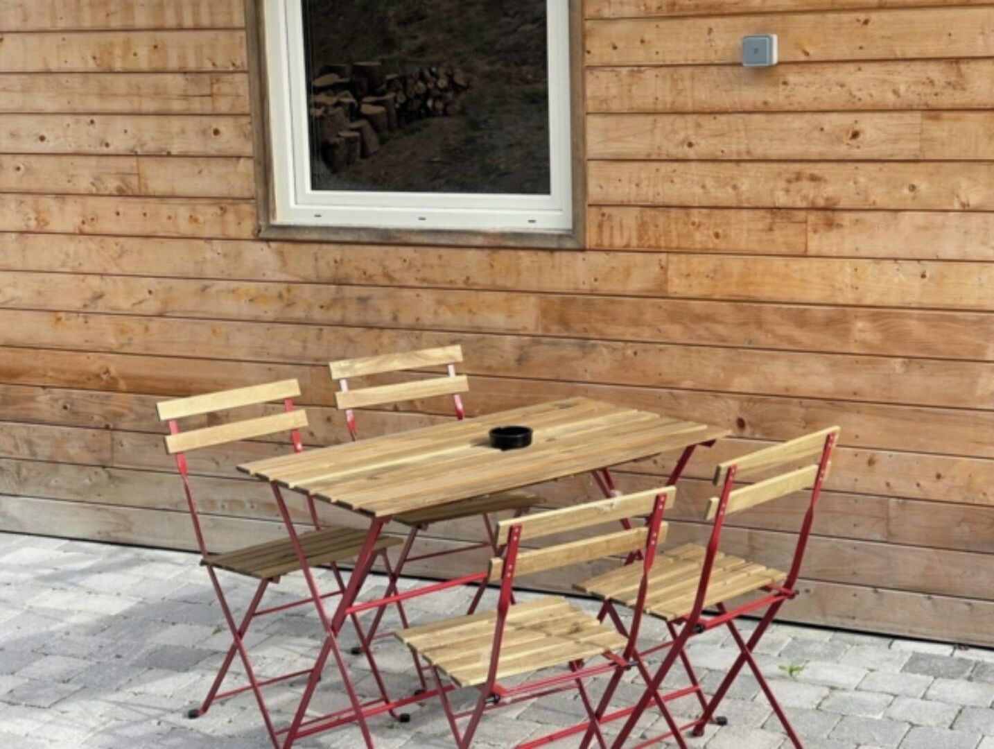 A wooden cabin with a dining table and chairs, a window with a view of stacked firewood, and a metal railing.