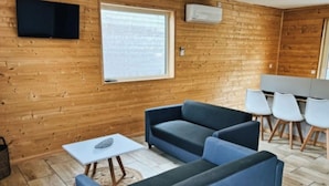 Ett rum med träpanel på väggarna, en platt-TV som hänger ovanför ett fönster, en soffa, ett litet bord och en sittgrupp med stolar.