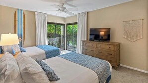 A hotel room with two beds, a ceiling fan, a TV, a wooden dresser, and a balcony with curtains.