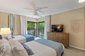 Habitación de hotel con dos camas, ventilador de techo, televisor, cómoda de madera y balcón con cortinas.