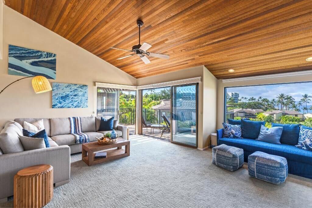 A spacious living room with a wooden ceiling, a large window offering a view of palm trees, and a blue sofa with matching cushions.