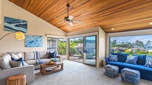 A spacious living room with a wooden ceiling, a large window offering a view of palm trees, and a blue sofa with matching cushions.