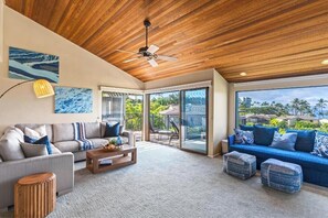 Ein geräumiges Wohnzimmer mit Holzbalkendecke, einem großen Fenster mit Blick auf Palmen und einem blauen Sofa mit passenden Kissen.