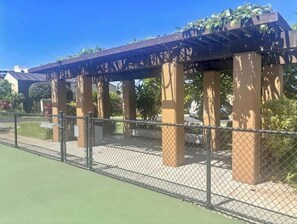 A covered outdoor seating area with a chain-link fence, concrete pillars, and a pergola with climbing plants. - Charming Oceanview Oasis: Wailea Elua 802 Surrounded by Lush Tropical Grounds (Wailea, Maui)