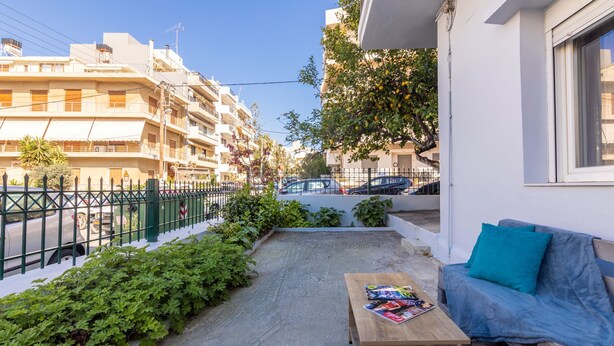 A balcony with a wooden table, a blue sofa, and a view of a residential street. - Klio's cozy house city center (Heraklion)
