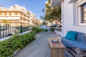 A balcony with a wooden table, a blue sofa, and a view of a residential street. - Klio's cozy house city center (Heraklion)