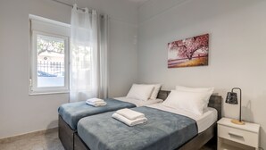 A bedroom with two beds, a window with curtains, a nightstand with a lamp, and a painting on the wall. - Klio's cozy house city center (Heraklion)