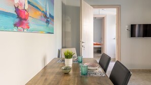 A dining area with a wooden table, chairs, a potted plant, and decorative items. A painting is on the wall, and a TV is mounted on the wall. - Klio's cozy house city center (Heraklion)