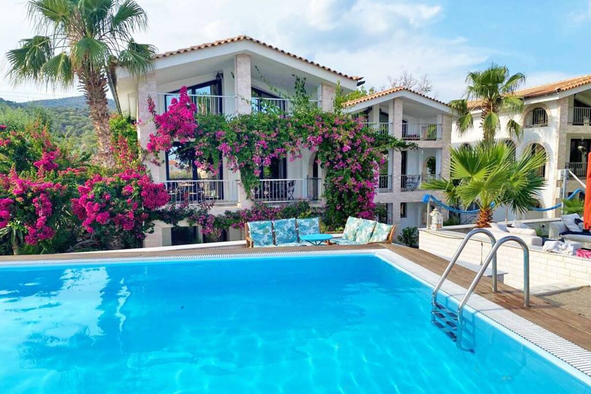 A swimming pool with a clear blue water, surrounded by a white building with balconies and pink flowering plants.