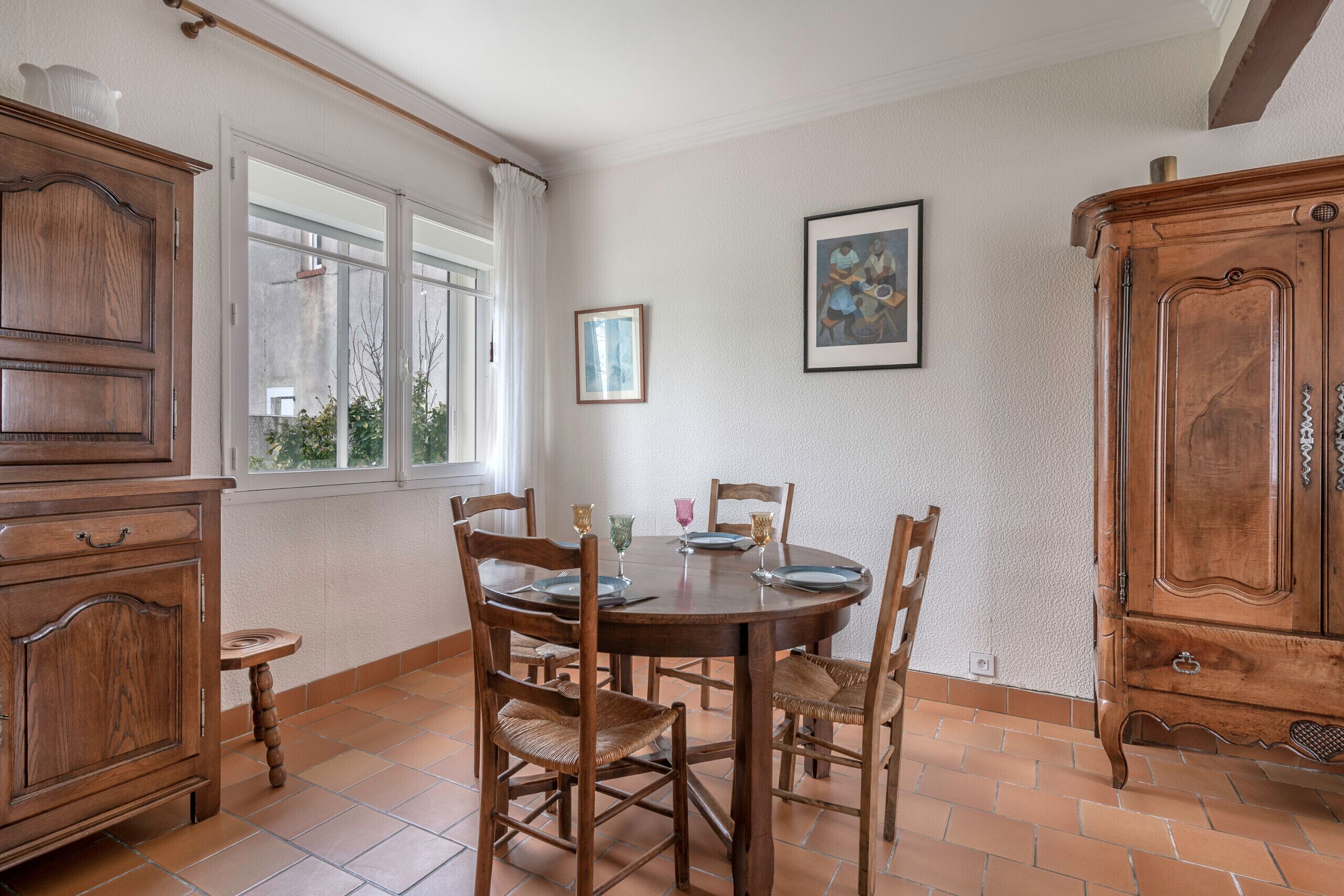 A dining area with a round wooden table, six chairs, and a window with curtains. There are two wooden cabinets and a framed picture on the wall.