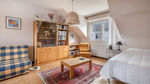Una habitación acogedora con un sofá a cuadros, una estantería de madera, un tapete estampado, una cama blanca y una ventana con cortinas.
