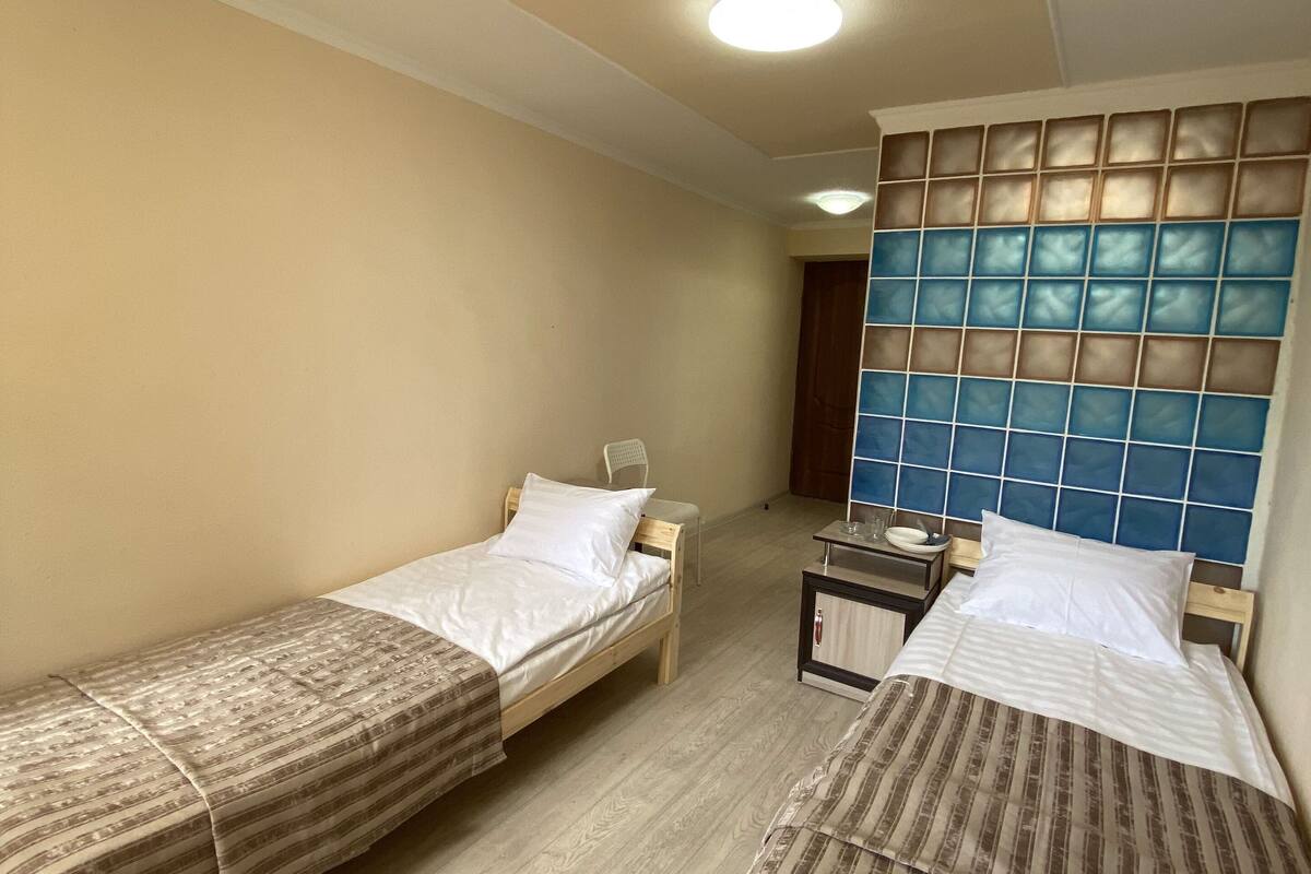 A room with two single beds, a small table, a chair, and a wall-mounted shelf.