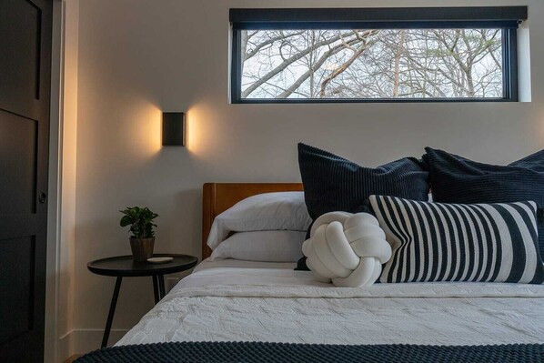 A bedroom with a bed, pillows, a bedside table with a plant, and a window with a view of bare trees.