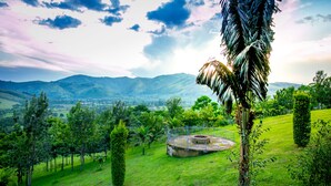 A lush green landscape with a circular stone structure surrounded by a metal fence, set against a backdrop of distant mountains and a partly cloudy sky. - THE CRESTED CRANE BWINDI HOTEL (Kyesero)