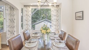 Une salle à manger avec une table dressée pour six personnes, un lustre et une vue sur les arbres par la fenêtre.