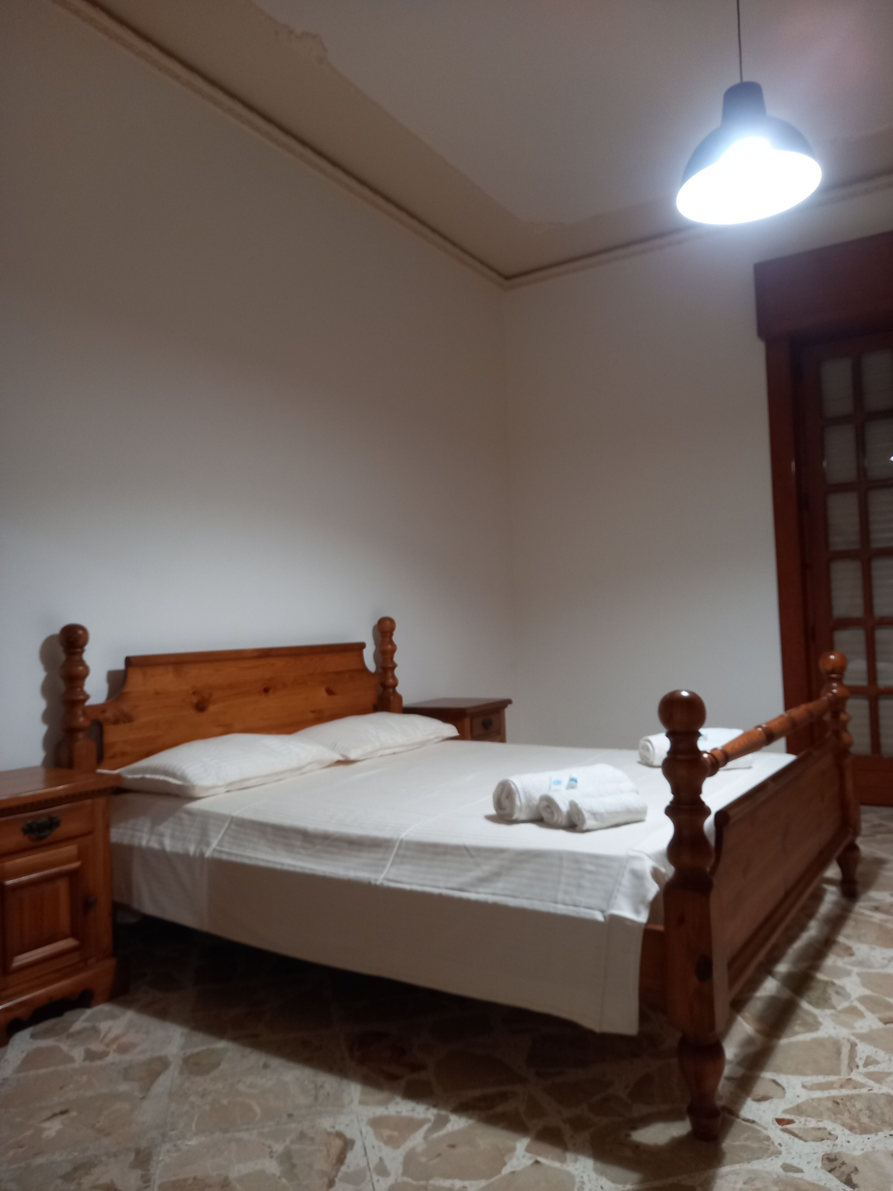 Una cama individual con cabecera y piecera de madera, una mesita de noche y un buró. Una lámpara colgante se encuentra sobre la cama.