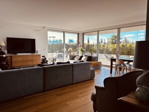 Un salon moderne doté d’un grand canapé d’angle gris, d’un téléviseur à écran plat, de planchers en bois et de grandes fenêtres offrant une vue sur l’extérieur.