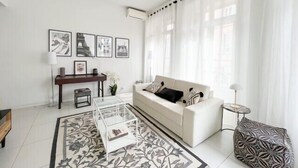 Ein modernes Wohnzimmer mit einem weißen Sofa, einem Glastisch und einem gemusterten Teppich. An den Wänden hängen gerahmte Bilder, und ein kleiner Beistelltisch sowie eine Lampe stehen bereit.