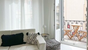 Ein Zimmer mit einem weißen Sofa, einem gemusterten Hocker und einem Balkon mit Holzbänken und Tischen.