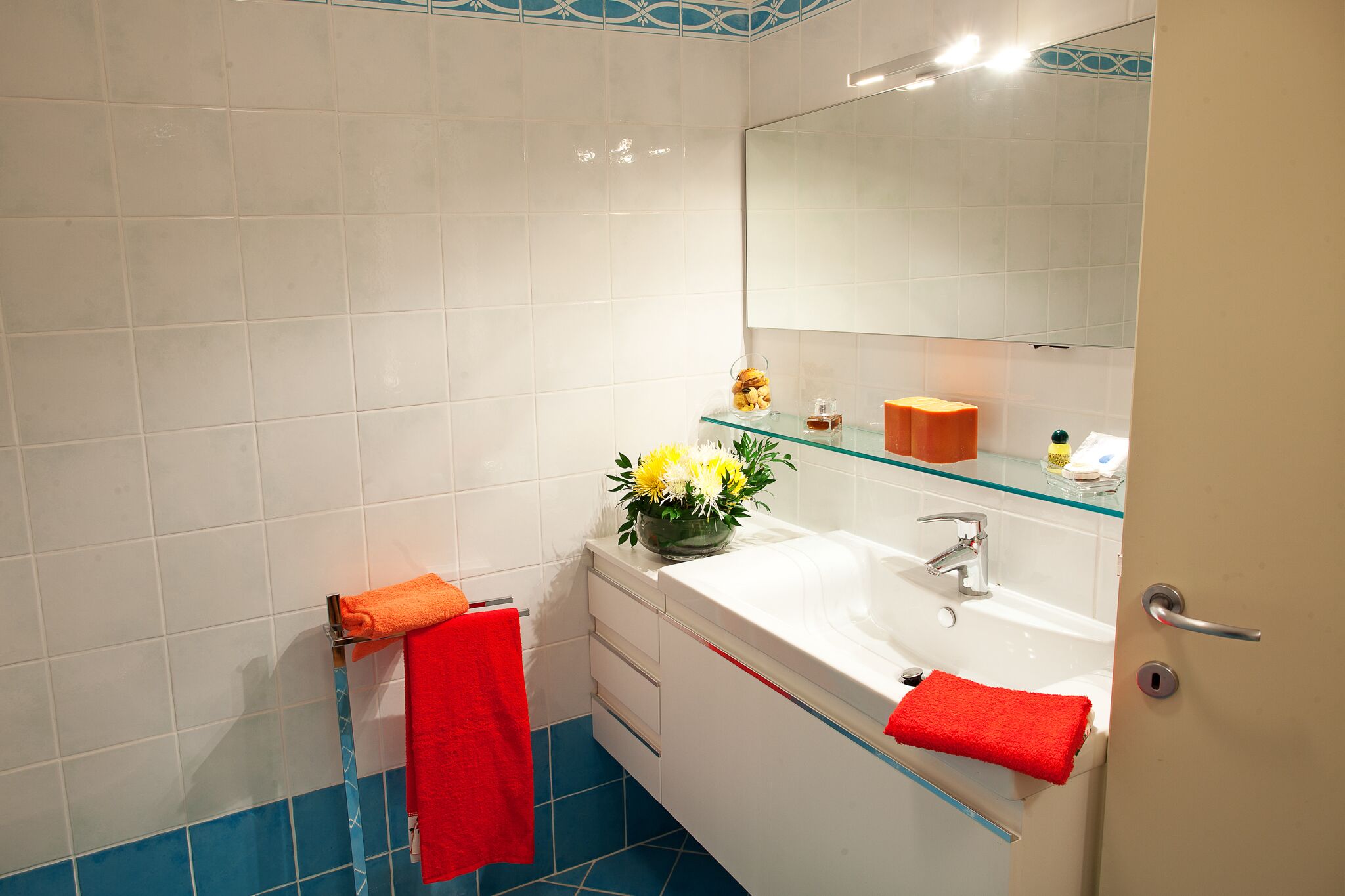 A bathroom with white tiles, a glass shelf with toiletries, a red towel, and a mirror.