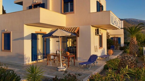 A modern two-story building with a patio area featuring a table and chairs, a sun umbrella, and a lounge chair. - 3-room House With Sea View (Ierapetra)