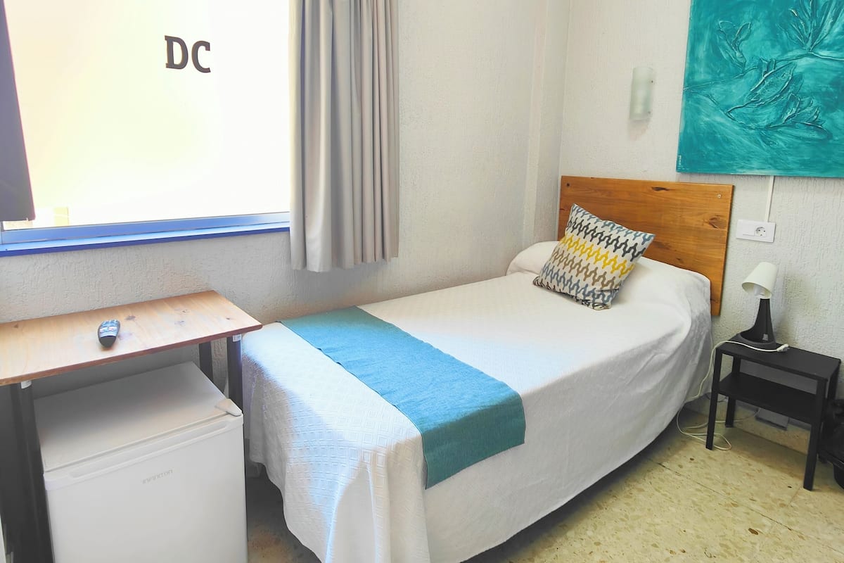 Una habitación pequeña y bien iluminada con una cama, un escritorio, un refrigerador pequeño y una ventana con cortinas.