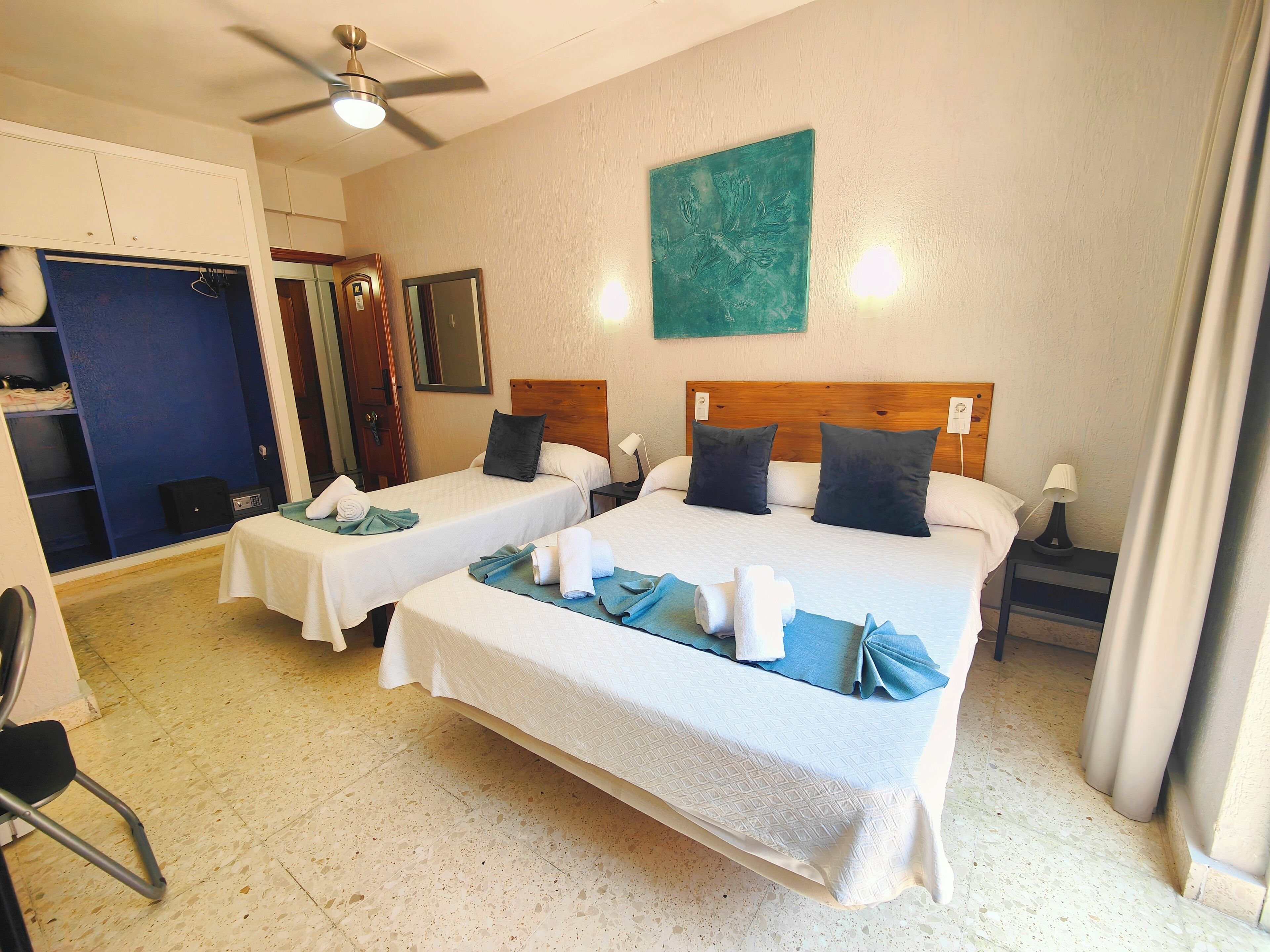 Habitación de hotel con dos camas, un ventilador de techo, un cuadro en la pared y una mesita con toallas.