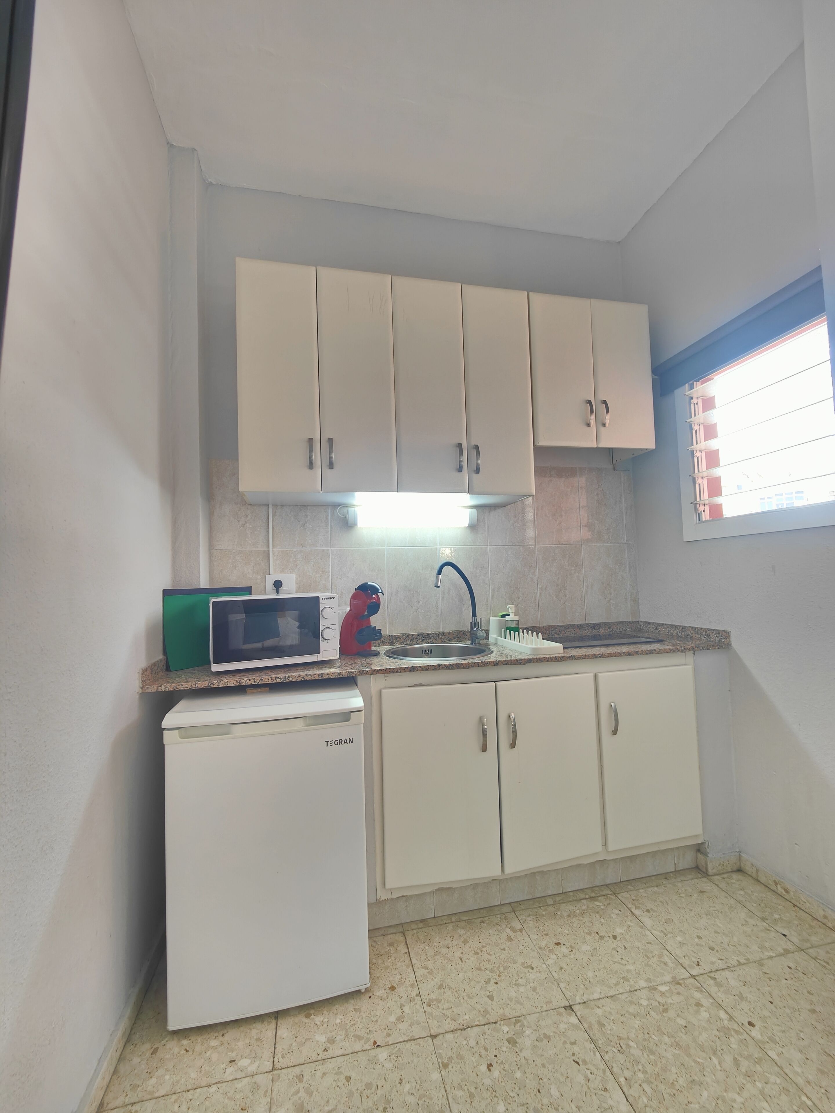 Una cocina compacta con armarios blancos, microondas integrado, un refrigerador pequeño y una ventana con persianas.