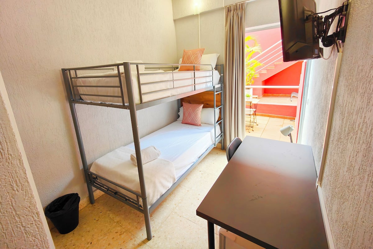 Habitación con literas, escritorio, una silla, un cubo de basura pequeño y vista a un área exterior con un muro rojo y una planta.