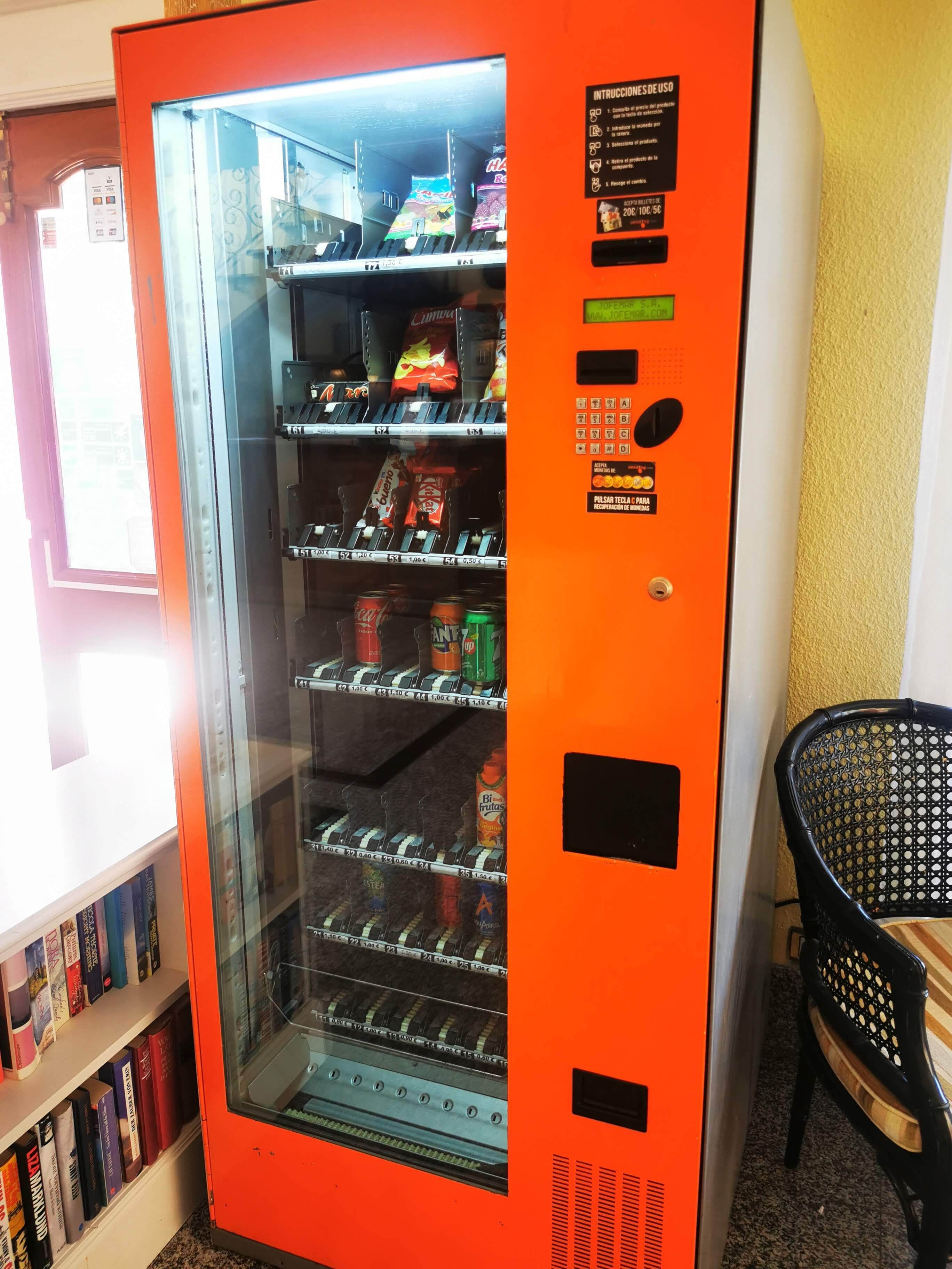 A vending machine stocked with snacks and drinks, located next to a bookshelf and a chair.