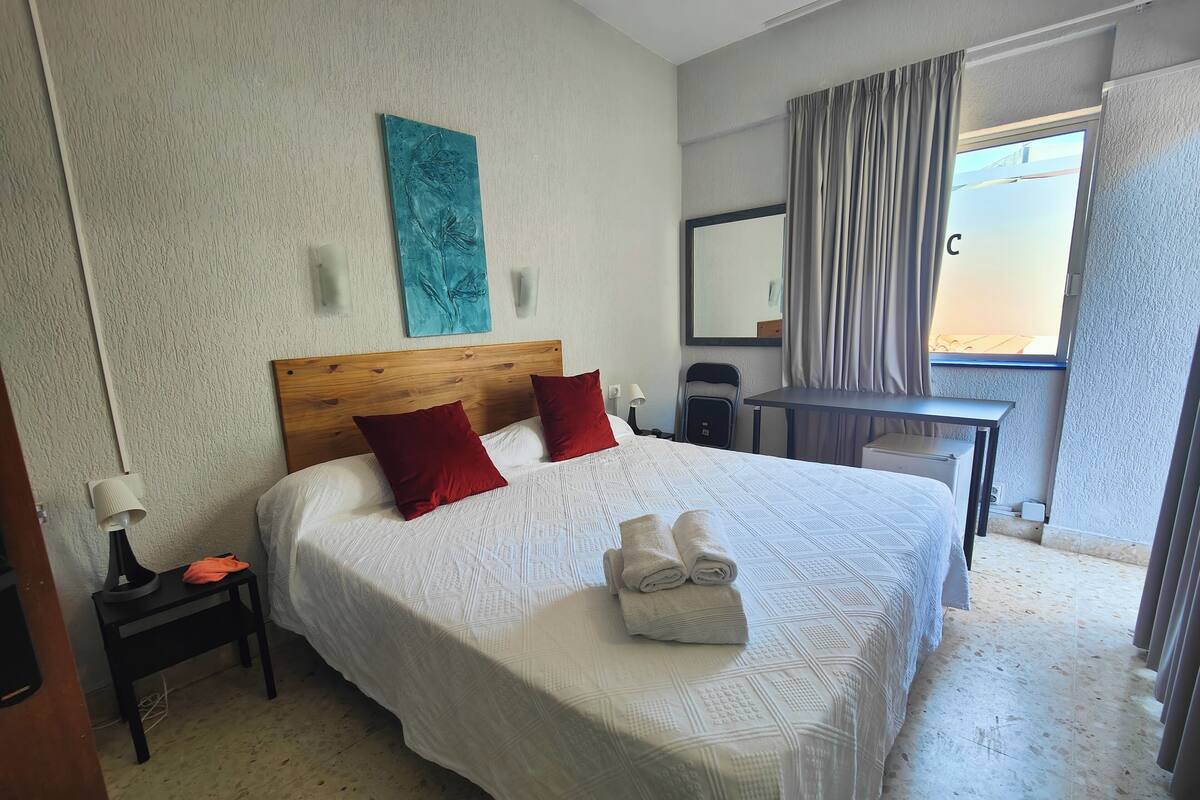 Habitación de hotel con una cama grande, dos almohadas rojas, un cabecero de madera, una obra de arte en la pared, un escritorio, una silla y una ventana con cortinas.