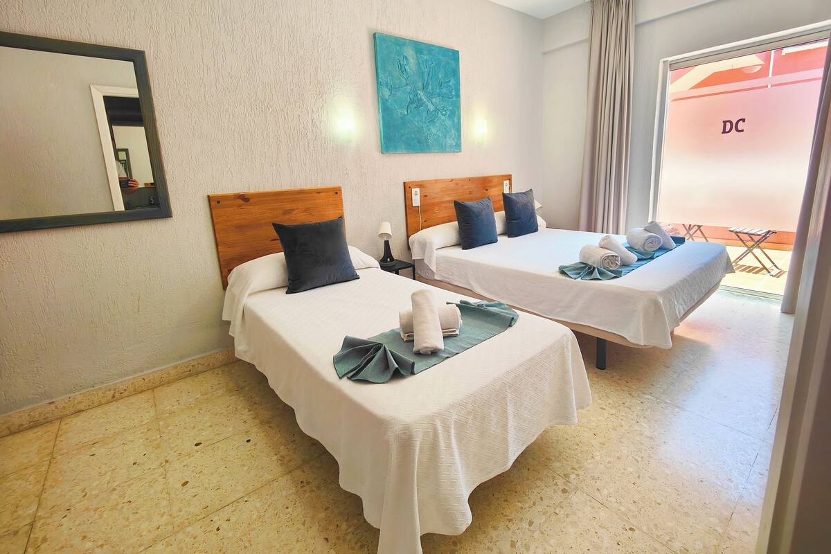 Habitación de hotel con dos camas individuales, un espejo, una obra de arte azul y un balcón con un letrero 'DC'.