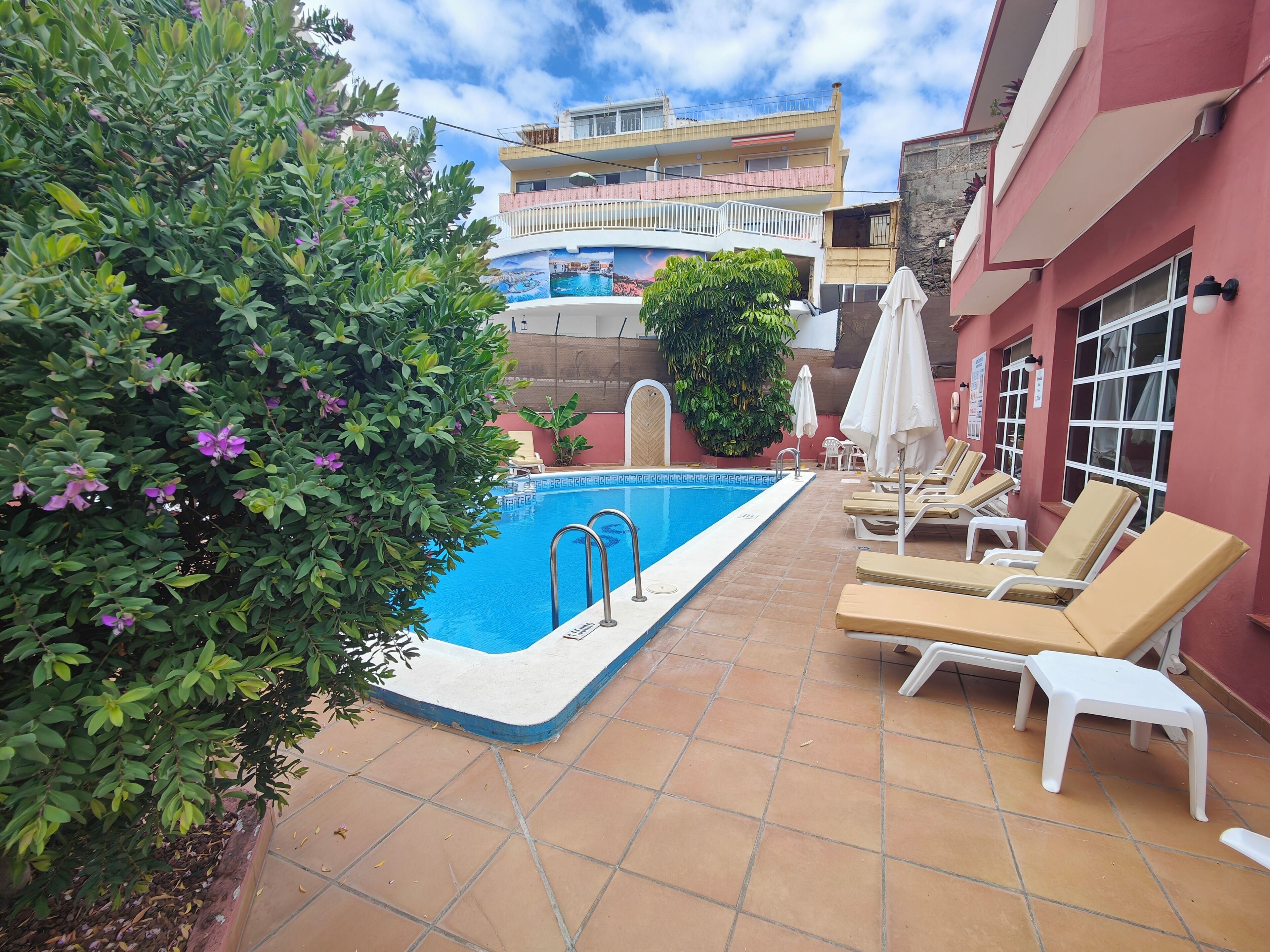A swimming pool with lounge chairs and umbrellas, surrounded by greenery and a building with balconies.