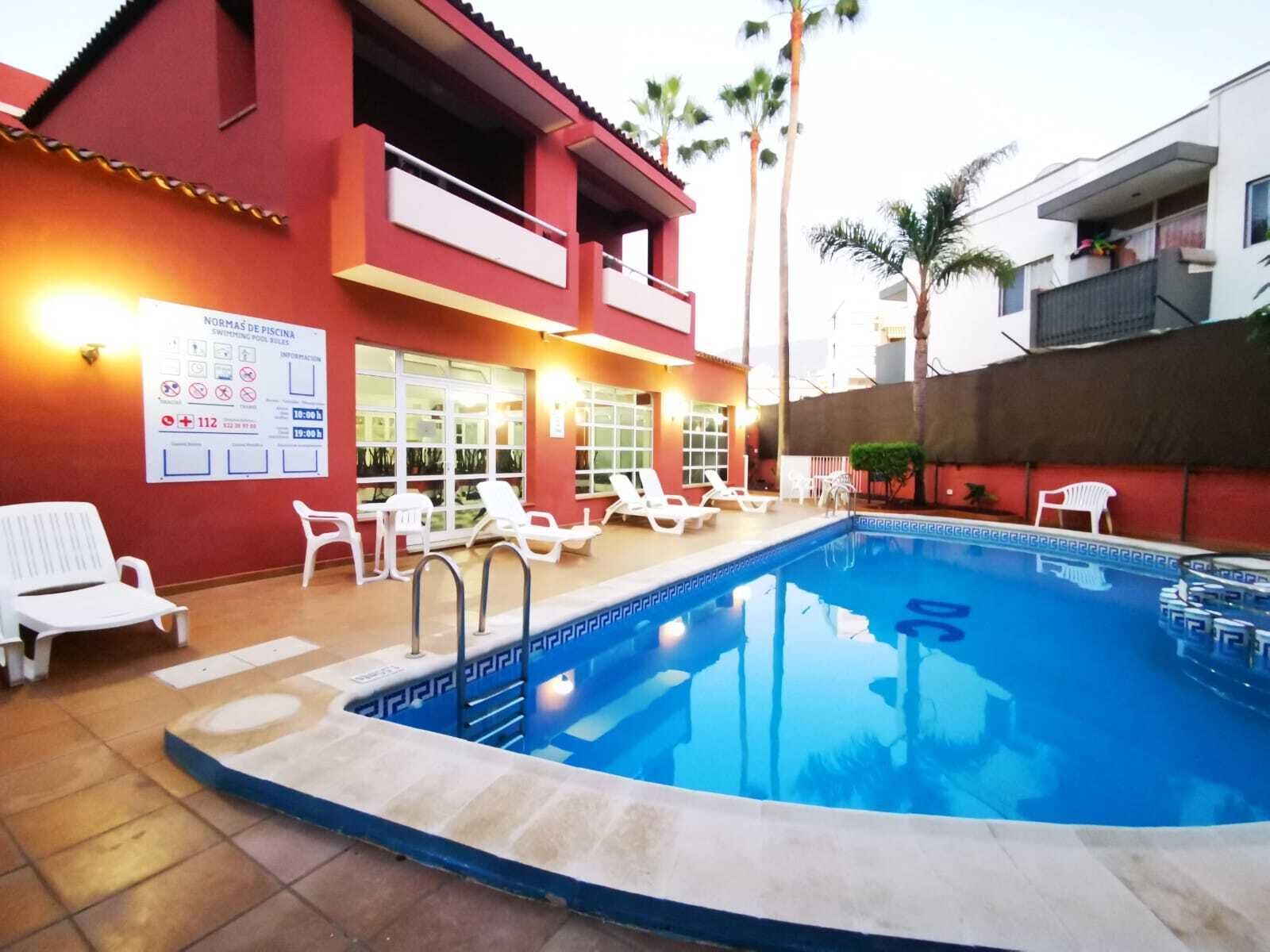 Zona de piscina con sillas de descanso y un edificio rojo con ventanas.