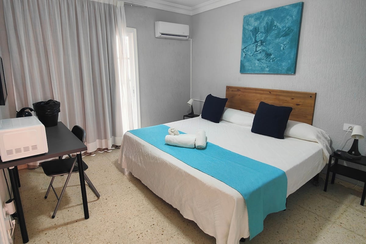 Habitación de hotel con cama, escritorio, silla, ventilador de techo, equipo de aire acondicionado y un cuadro en la pared.