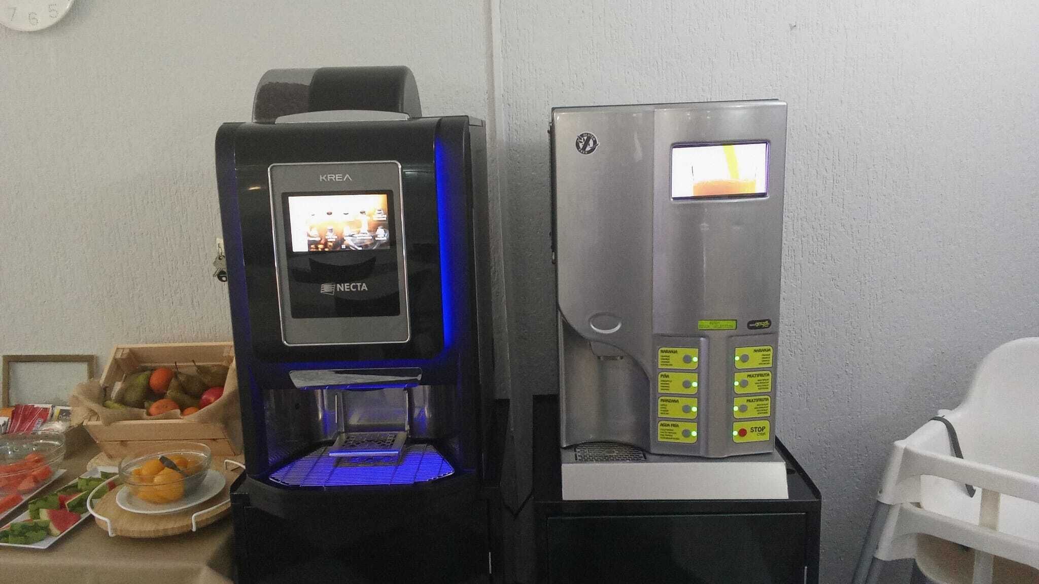 Deux distributeurs de boissons, l’un avec un éclairage bleu et l’autre avec un écran numérique, sont placés à côté d’un saladier rempli de fruits et d’une chaise blanche.