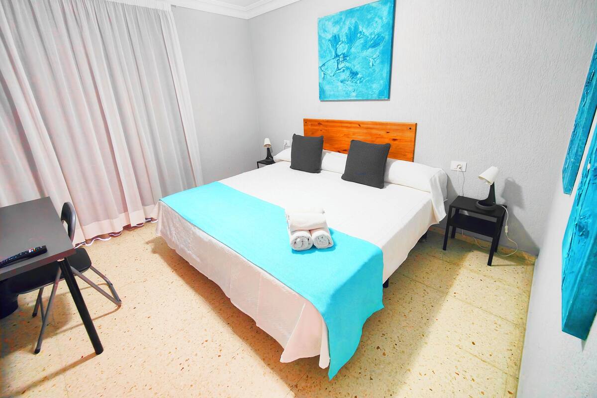 Una habitación de hotel con cama, escritorio, silla, mesita de noche, lámpara y un cuadro en la pared.
