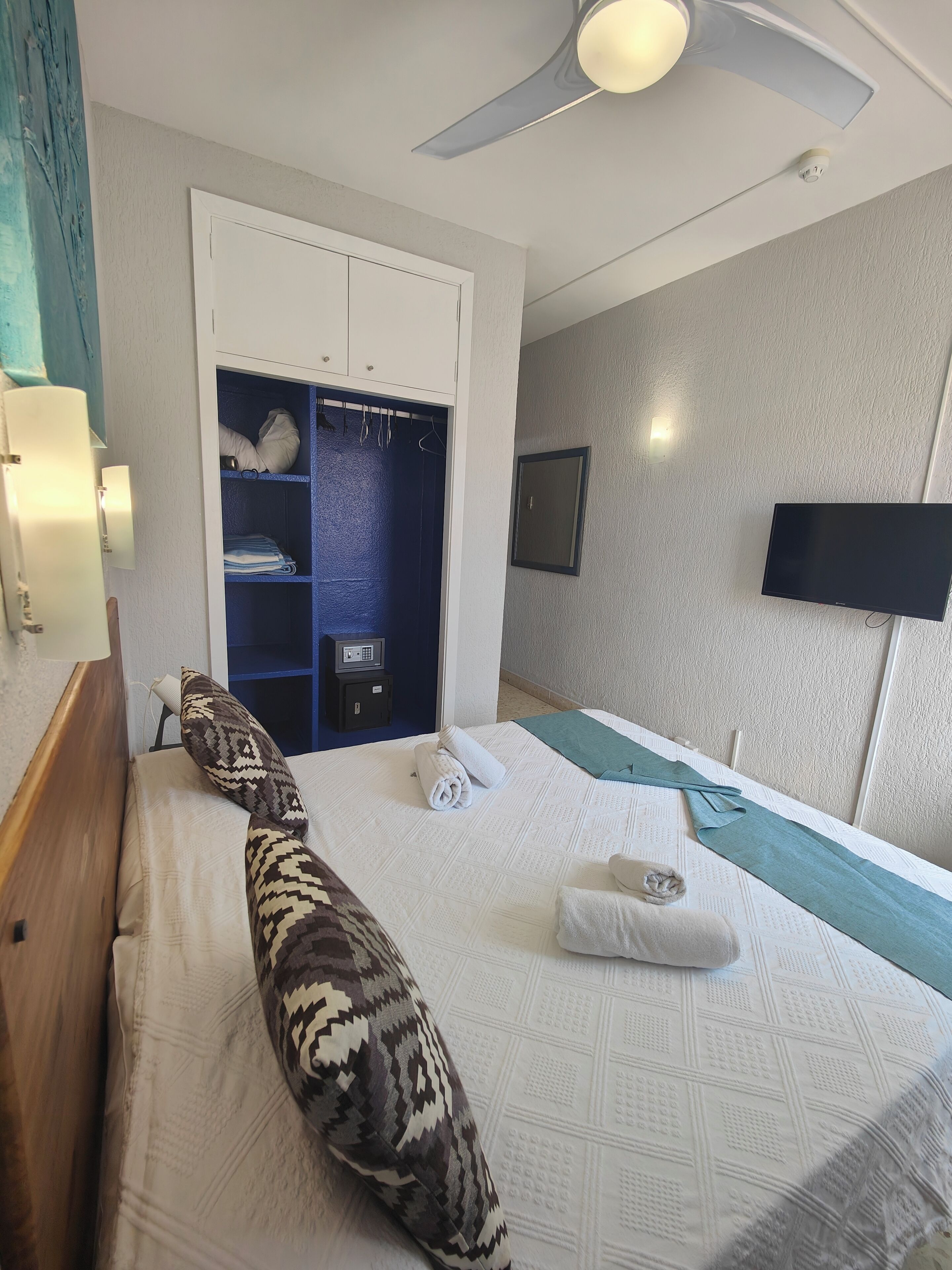 Una habitación de hotel con cama, armario, televisor de pantalla plana y ventilador de techo.