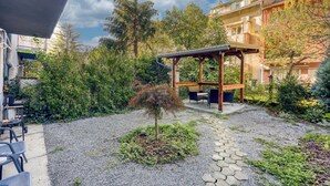 Een overdekt terras met een pergola, omgeven door een tuin met een kleine boom en grindpaden.
