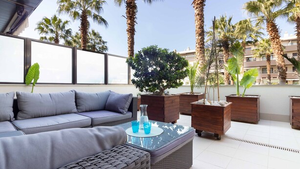 A rooftop terrace with a grey sofa, wicker ottoman, and potted plants.