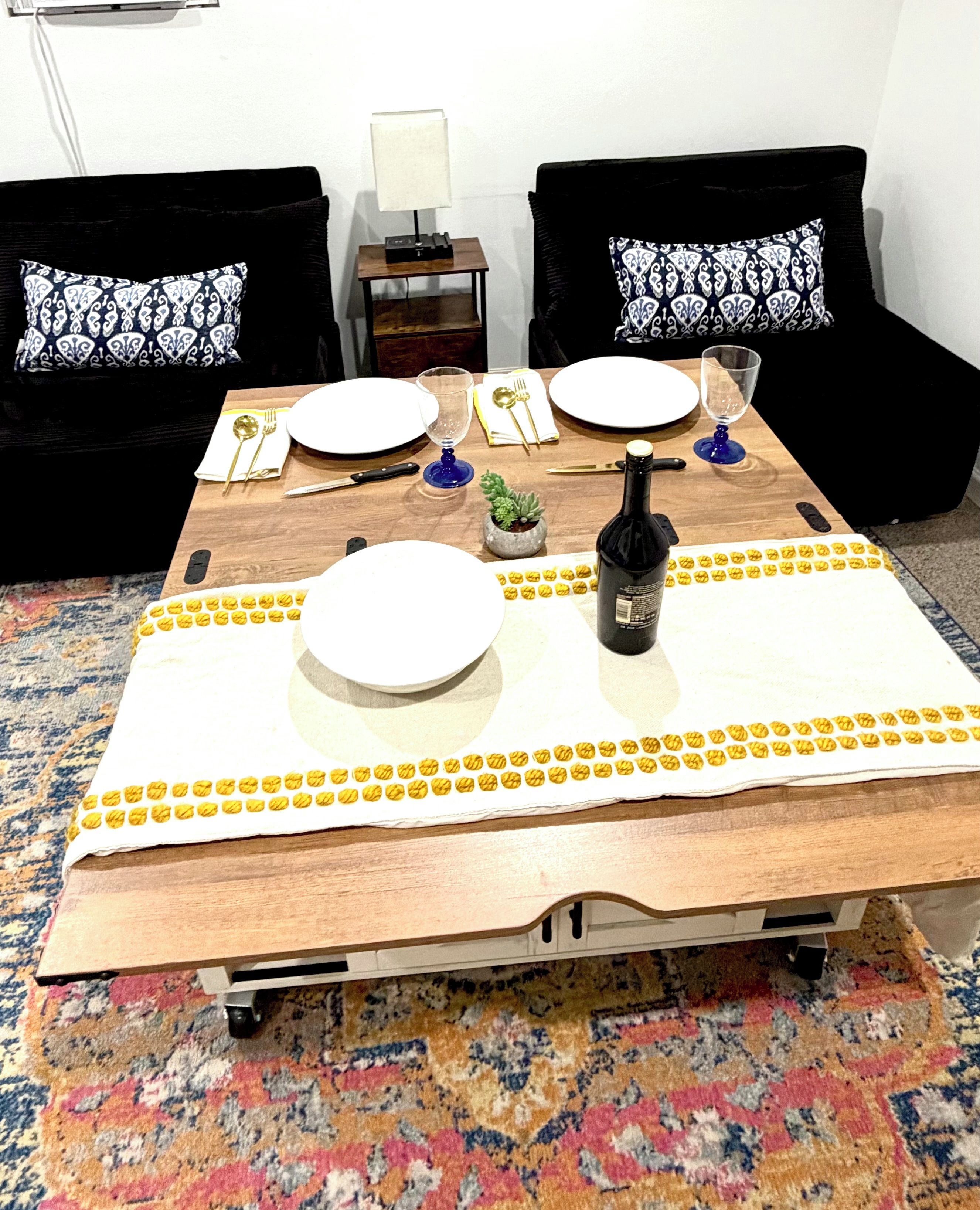 A dining table set with plates, cutlery, and a bottle, surrounded by black sofas with patterned cushions.