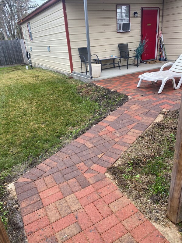 Un chemin de briques mène à une maison avec une porte rouge, une petite terrasse avec des chaises et une cour clôturée.