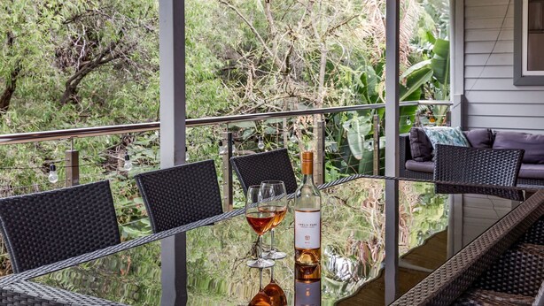 A glass table with chairs, a bottle of wine, and two wine glasses on a balcony with a view of greenery.