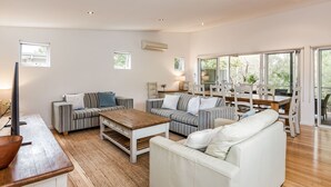 A spacious living area with a sofa, a coffee table, and a dining table. There are multiple windows providing natural light and a view of the outdoors.