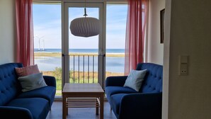 A room with a balcony overlooking a beach and wind turbines, featuring two blue sofas and a hanging light fixture. - Large holiday house with great seaview - sauna and hottub (Glesborg, Bønnerup Strand)